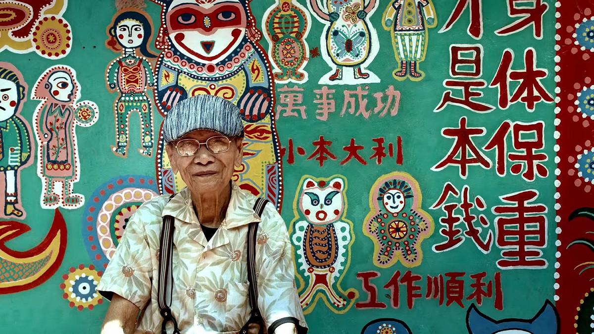 花博拍攝日文形象影片中出現「彩虹爺爺」黃永阜，以佈滿皺紋的手，執筆繪出知名的旅遊景點「彩虹眷村」，飽和的鮮艷色彩、生動逗趣的人物與動物。 圖：台中市政府提供