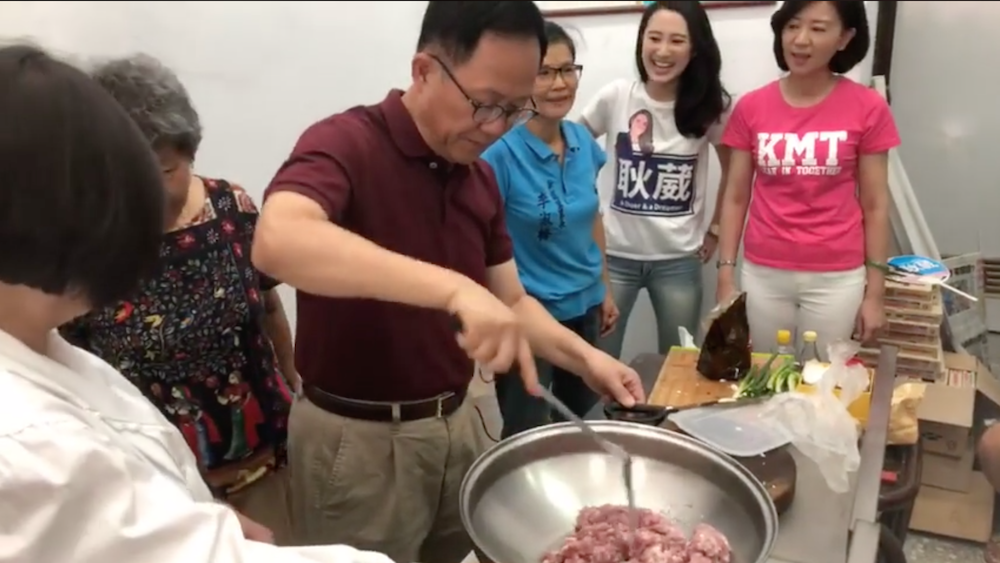 國民黨台北市長參選人丁守中上午到新龍里煮麻婆豆腐給里民老人吃，由於室內悶熱，幾位媽媽們站在丁守中後面貼心的幫丁守中搧扇子，但立刻被一旁的國民黨台北市黨部主委黃茹錦（左）斥喝：「怕熱就不要進廚房！」要求媽媽們不要為丁守中搧扇子！ 圖：翻攝自丁守中臉書