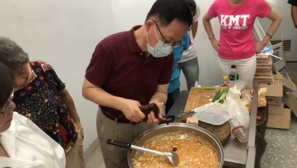 國民黨台北市長參選人丁守中上午到新龍里煮麻婆豆腐給里民老人吃，由於室內悶熱，幾位媽媽們站在丁守中後面貼心的幫丁守中搧扇子，但立刻被一旁的國民黨台北市黨部主委黃茹錦（左）斥喝：「怕熱就不要進廚房！」要求媽媽們不要為丁守中搧扇子！ 圖：翻攝自丁守中臉書