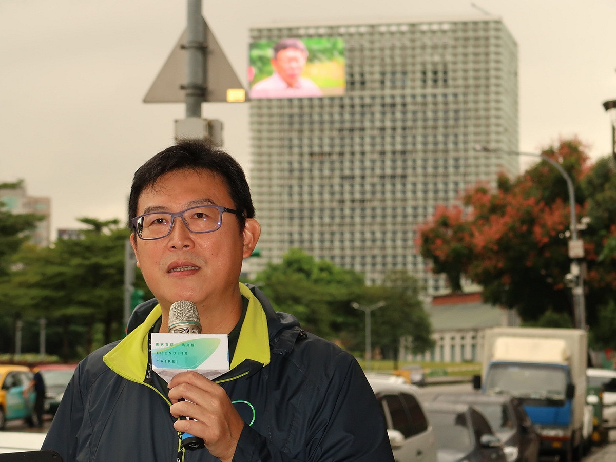 民進黨台北市長候選人姚文智17日在三創園區遠方召開記者會，而背後LED廣告牆正播放柯文哲的市府廣告。 圖：林朝億/攝