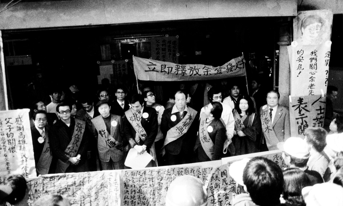 自左一起為郭一成、張俊宏、許信良、姚嘉文、黃順興、周平德、施明德、陳菊、何春木等人，個個身披紅色布條，書寫著自己的名字在橋頭余家門口，高舉「立即釋放余登發父子」前布條合影。攝影/陳博文 圖：陳博文／攝