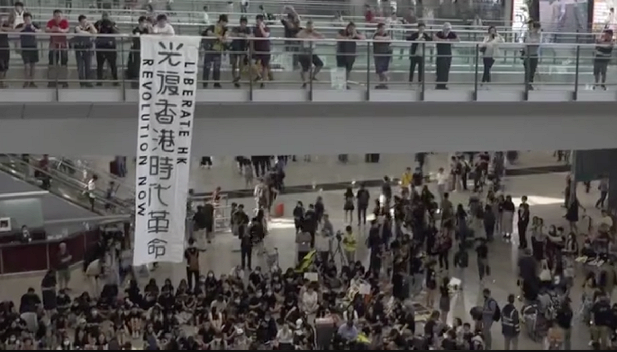 香港網友發起「萬人接機」集會，引起逾千人參與，香港機場的1號客運大樓的行人橋上，有人掛出「光復香港 時代革命」標語，吸引不少旅客注意。 圖：翻攝自港媒《立場新聞》直播