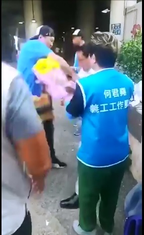 何君堯在屯門設攤，1名藍衣男子（左）獻上太陽花後突然抽刀砍向何君堯（右二白衣者）。 圖：翻攝自香港電台視像新聞臉書影片