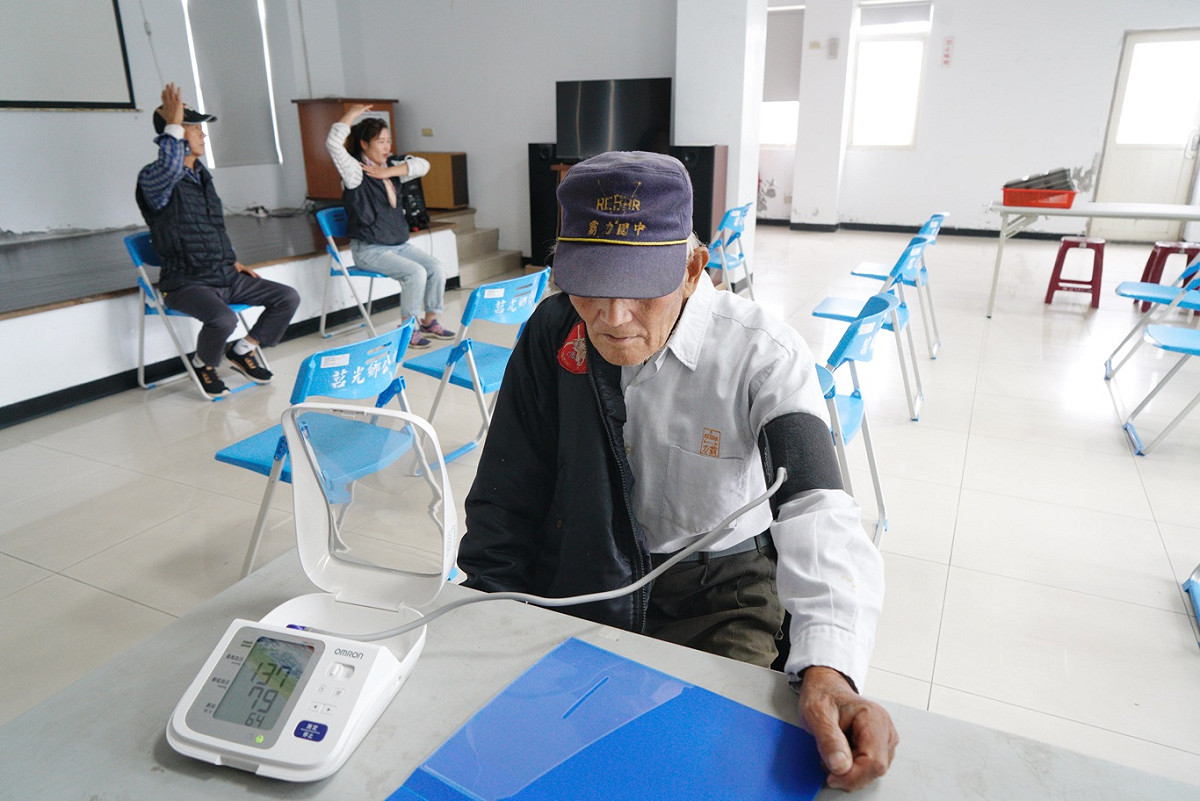 東莒一位長者出席老人共餐活動，島上衛生所的工作人員幫他量血壓，兩位居服員則正為帶動老人的肢體運動彩排。 圖：張良一/攝