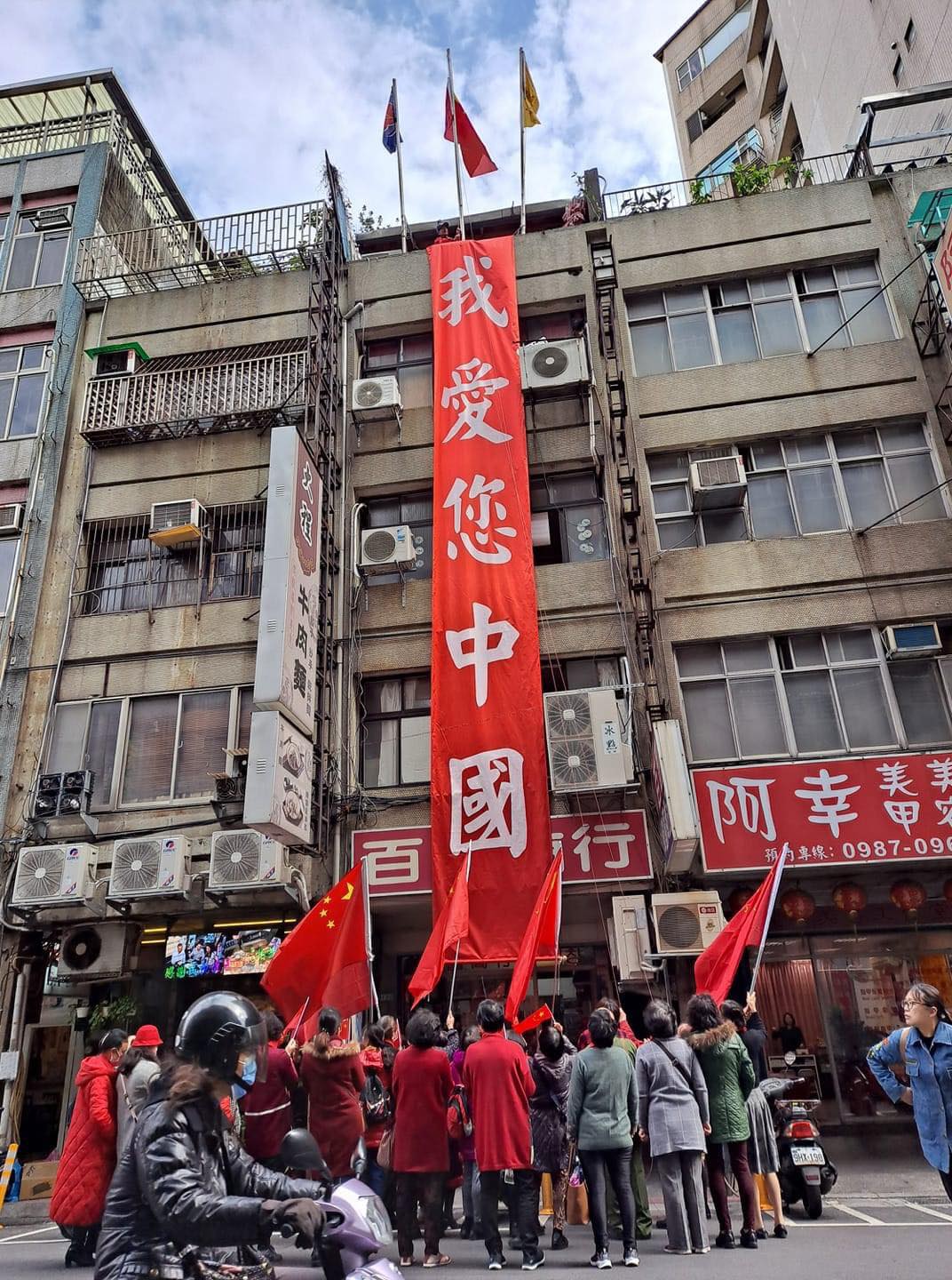 台北市街頭某處大樓外牆，卻掛上大大的紅布條，上頭寫著「我愛您中國」。1樓還有一群人高舉著「中國五星旗」 圖：翻攝自 鄉民挺起來 臉書