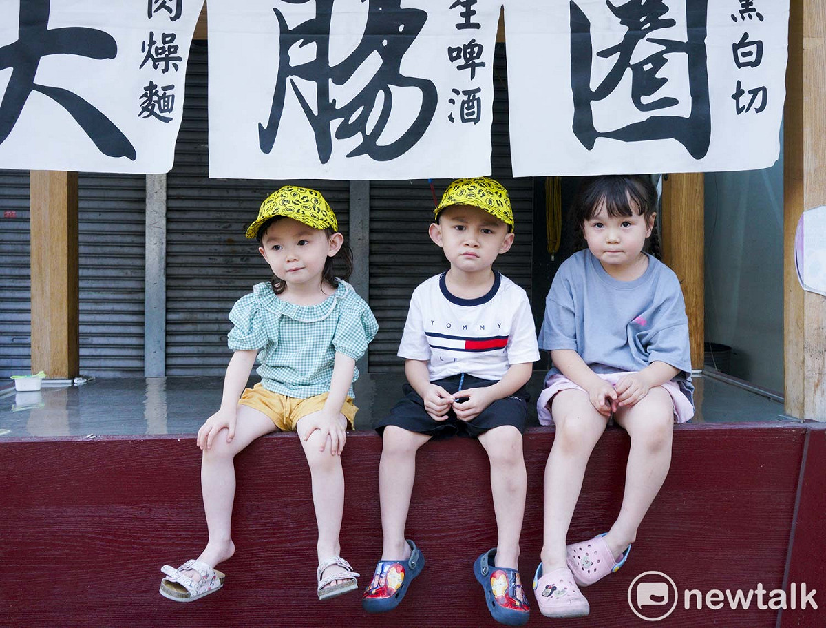 三位小朋友坐在彰化市中山路上賣大腸圈的攤子上，看著白沙屯媽祖往北港朝天宮進香的盛況。 圖：張良一/攝