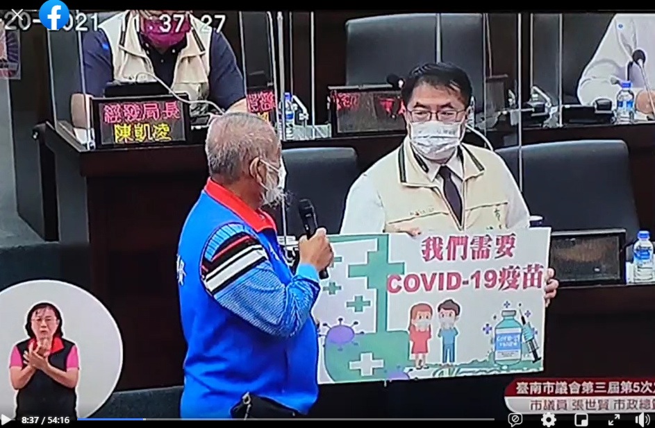 張世賢拿著｢我們需要covid-19疫苗｣紙板到備詢台，要求黃偉哲簽名支持。 圖：翻攝張世賢臉書