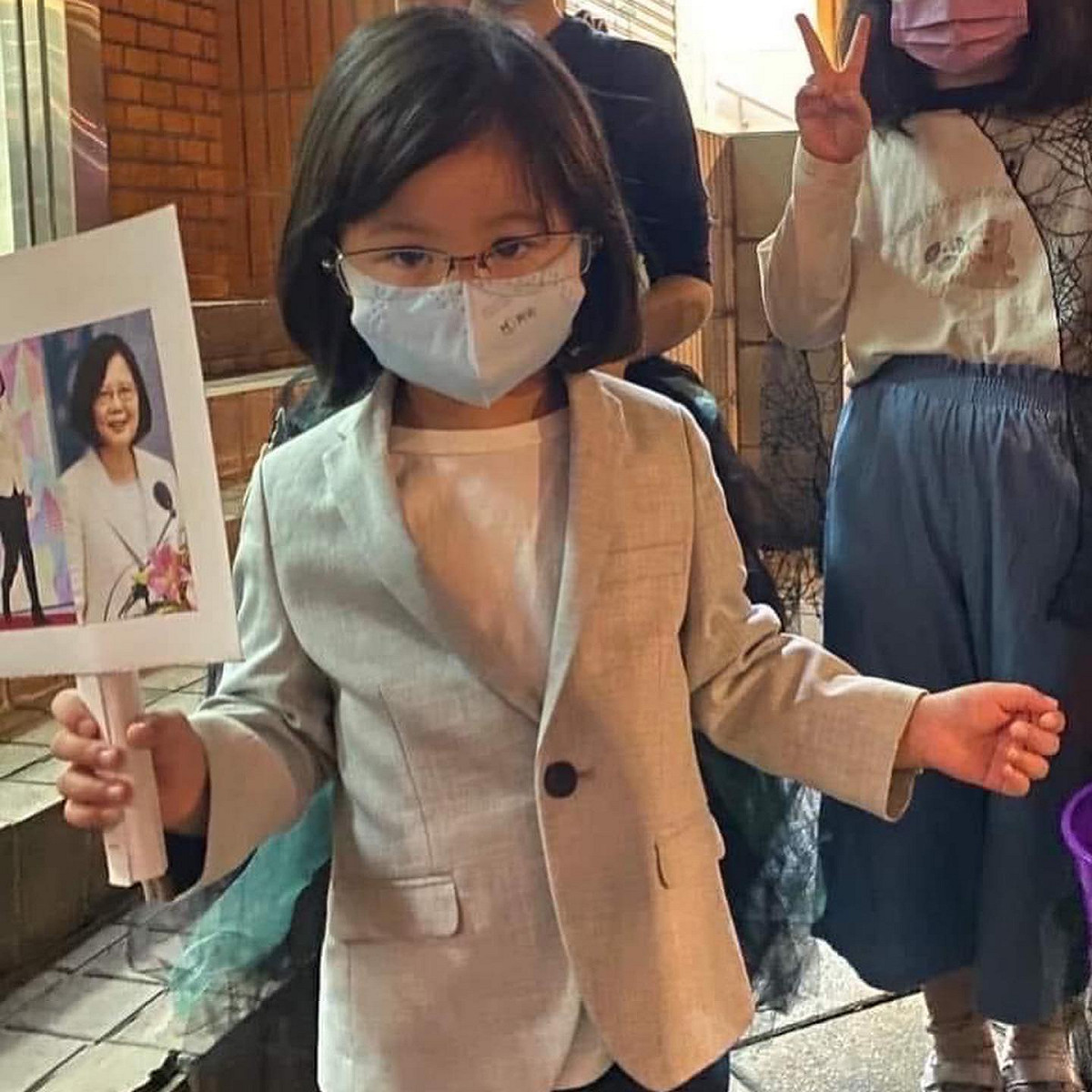 「迷你小英」戴上假髮和眼鏡，穿上與總統幾乎同款的灰西裝、白衣、黑褲，並舉著一張貼有蔡英文照片的牌子，和總統相似度百分百。 圖：擷取自「Gangs of Tian Mu 天母幫」臉書社團