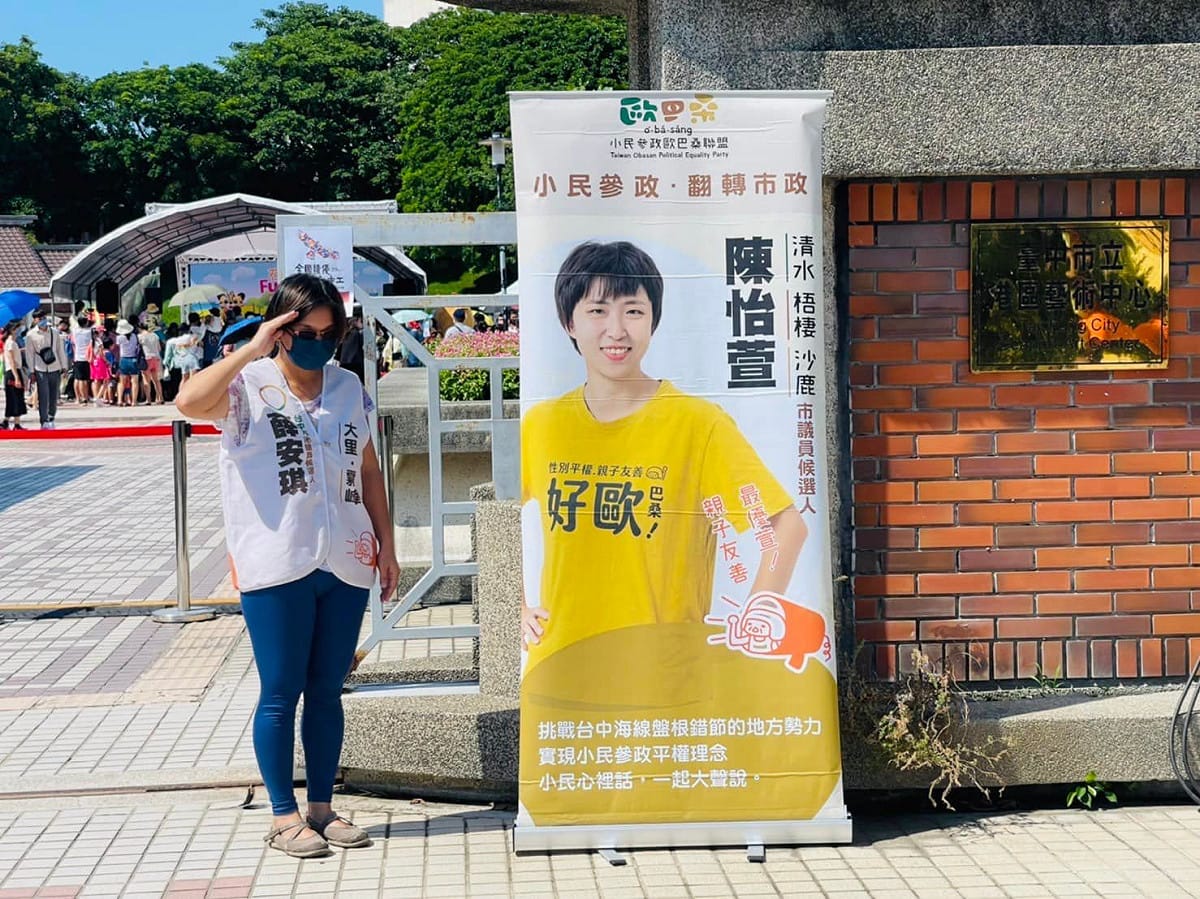 薛安琪在活動會樹下乘涼被趕，只能帶著陳怡萱的宣傳旗到門口拍照。  取自薛安琪臉書