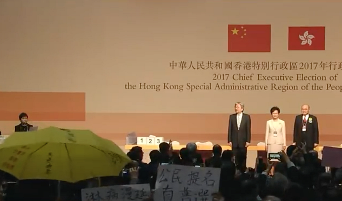 林鄭月娥、曾俊華與胡國興3名候選人在台上聆聽選舉結果，台下民眾高舉雨傘、「我要真普選」標語抗議。 圖：翻攝自香港蘋果日報直播畫面