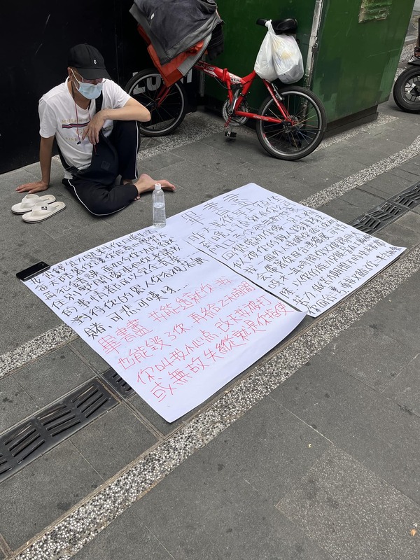 一名男子坐在路邊擺大字報，痛批畢書盡「功成名就後就對恩人視若無睹」。 圖：翻攝自路上觀察學院FB