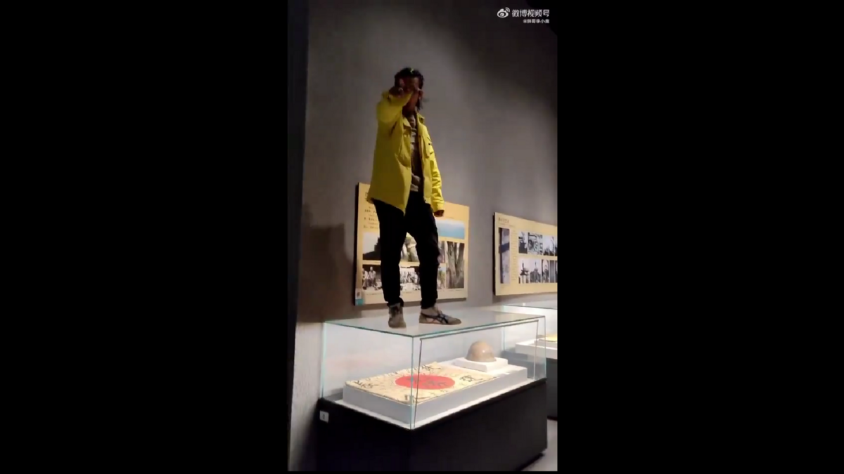 一名穿著黃色衣服的男子在博物館內，跳上了展示櫃，櫃中擺放著疑似二戰時期日軍用品，並做出勝利的手勢。 圖:翻攝自影片