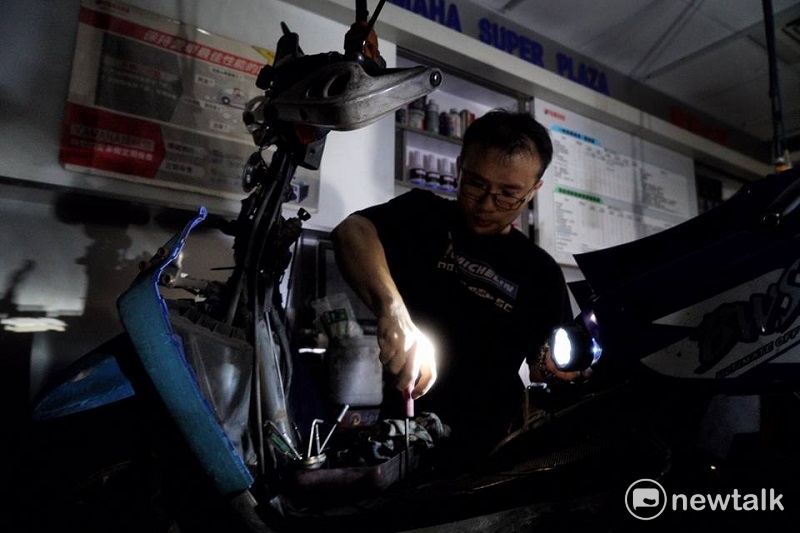 一家機車行老闆為減少損失，只好一手持手電筒，一手修車。 圖：常日豪/攝