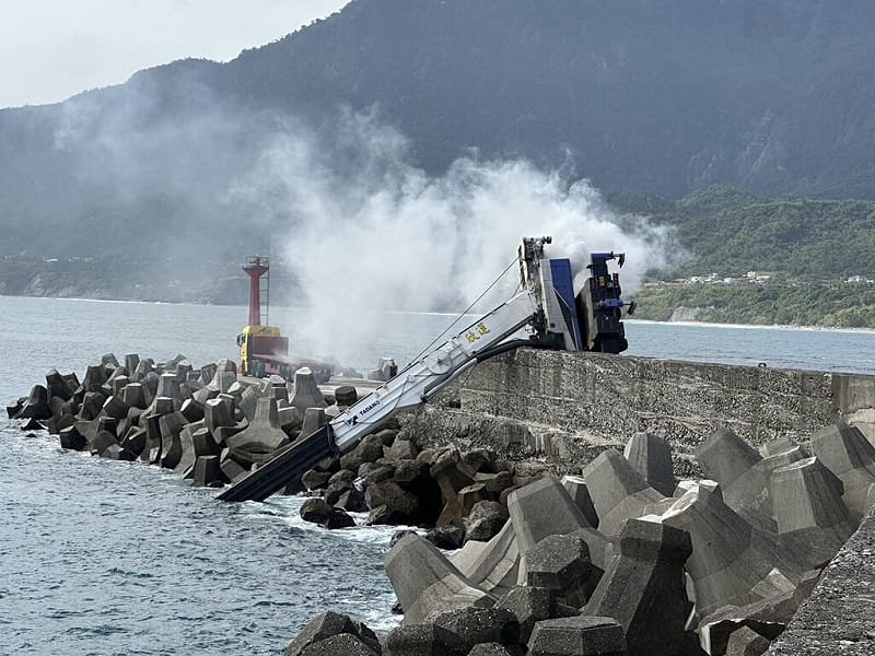 台東成功漁港30日中午一輛吊車在碼頭吊掛消波塊時翻覆，車上其中1人在駕駛座已明顯死亡，另1人腿部遭車體壓住受困，消防人員救出後送醫救治，吊車還冒煙起火，隨即被警消撲滅。（民眾提供）中央社記者李先鳳傳真　114年11月30日   
