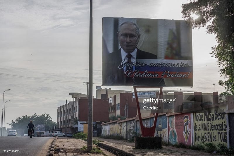 馬利首都巴馬科街頭仍可見俄羅斯總統普丁的巨型宣傳看板，上方寫著「生日快樂—弗拉基米爾·普丁」，而附近牆面則出現以黑字書寫的標語，呼籲「真正的民主與全民福祉」，凸顯當地政治立場分裂與社會張力升高。 圖:翻攝自X