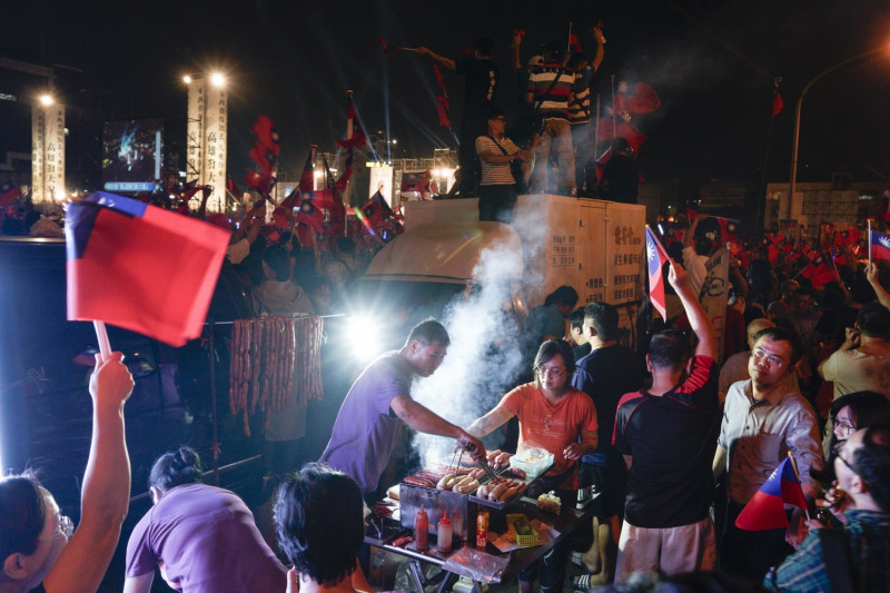 韓國瑜支持者在鳳山造勢晚會會場外的香腸攤等著買香腸，後頭的支持者則是激動的揮舞旗幟呼應舞台上的講者。   圖：張良一/攝
