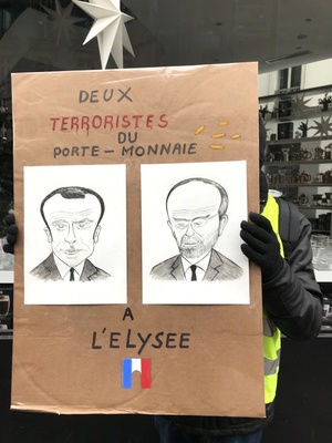 法國「黃背心」運動進行第5波抗議，一名抗議者舉著漫畫看板，上面有總統馬克宏與總理菲力普的肖像，一旁寫著「兩名（總統府）艾里賽宮的錢包恐怖份子」。   圖／中央社