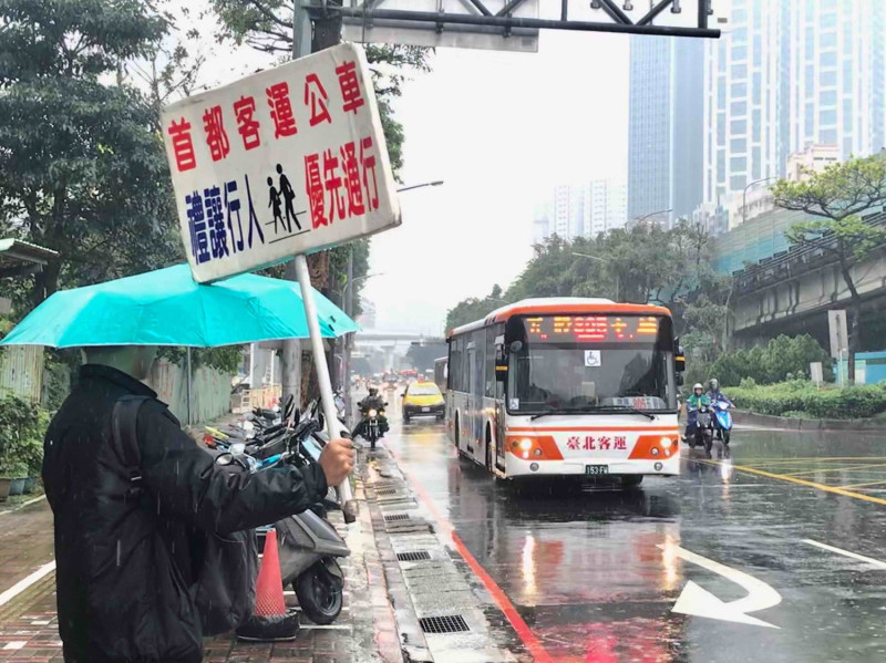 今早雖下雨，首都客運仍派員在公車進入路口前，舉牌向公車駕駛宣導「禮讓行人」。    圖：新北市交通局提供