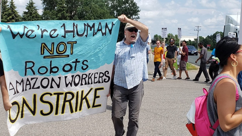 美國電商巨擘亞馬遜明尼蘇達州的配送中心員工在廠房外罷工，揮舞著「我們是人，不是機器人」的標語。   圖：翻攝Awood Center臉書