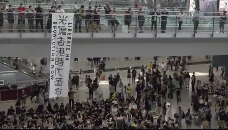 香港網友發起「萬人接機」集會，引起逾千人參與，香港機場的1號客運大樓的行人橋上，有人掛出「光復香港 時代革命」標語，吸引不少旅客注意。   圖：翻攝自港媒《立場新聞》直播