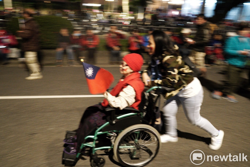 年輕人推著戴紅帽、穿紅背心且行動不便的婦人參加韓國瑜的造勢活動。   圖：張良一/攝