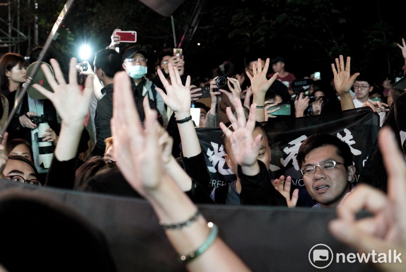 港人在「光復香港時代革命」旗幟後，以不曝光方式比出五大訴求手勢。   圖：張良一/攝