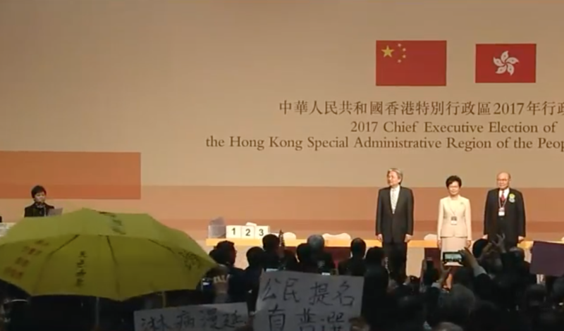 林鄭月娥、曾俊華與胡國興3名候選人在台上聆聽選舉結果，台下民眾高舉雨傘、「我要真普選」標語抗議。   圖：翻攝自香港蘋果日報直播畫面