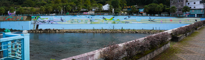 坪林區北勢溪茶博館前美化後的護岸彩繪，在北勢溪清澈的溪水映射及純白色調的茶博館襯托下，整幅壁畫彷彿如躍然紙上的畫。   圖：新北市水利局提供