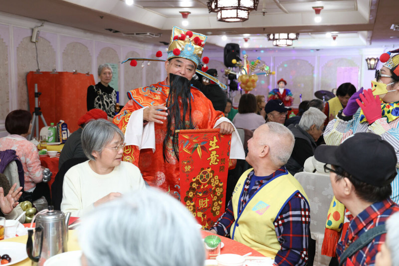 天主教失智老人基金會承辦之臺北市萬華老人服務中心舉辦「萬華獨居長者圍爐春節圍爐送暖餐會」，現場「財神爺」分送紅包，將溫暖與祝福傳遞給在場的每一位長輩。   圖:東森慈善基金會提供