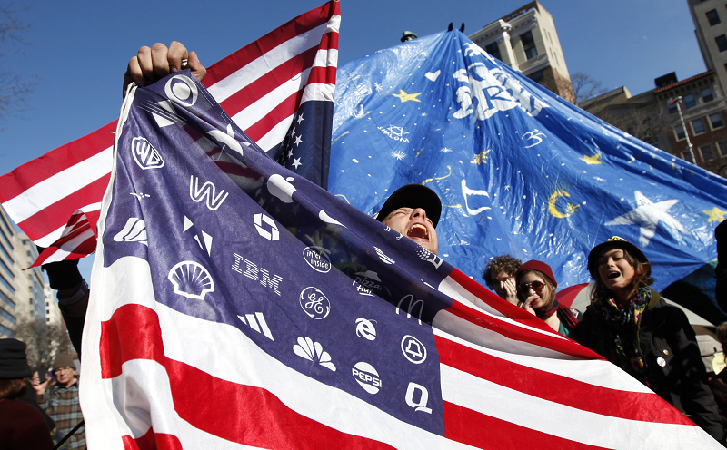 一名參與「佔領華府」（Occupy DC）的民眾手持特製的美國國旗，上頭的星星都被大企業的企業符號取代，諷刺財團介入國家的程度之高，他們身後則是佔領人架設的大型「夢想帳篷」。圖片來源：達志影像/路透社   