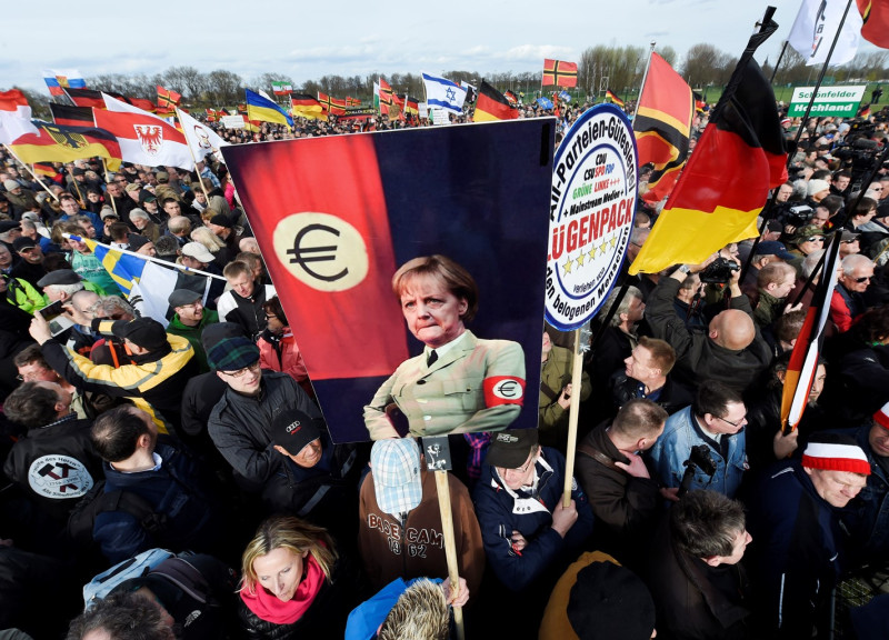 德國極右派團體「愛歐人反歐洲國家伊斯蘭化」（ PEGIDA）2015/4/13在東部德烈斯登遊行。示威者高舉穿著第三帝國制服，但以歐盟符號取代納粹的總理梅克爾標語。圖：達志影像/美聯社   
